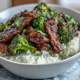 A close-up of a homemade Beef and Broccoli Bowl reveals glossy sauce coating beef and green broccoli, garnished with sesame seeds and green onions.  